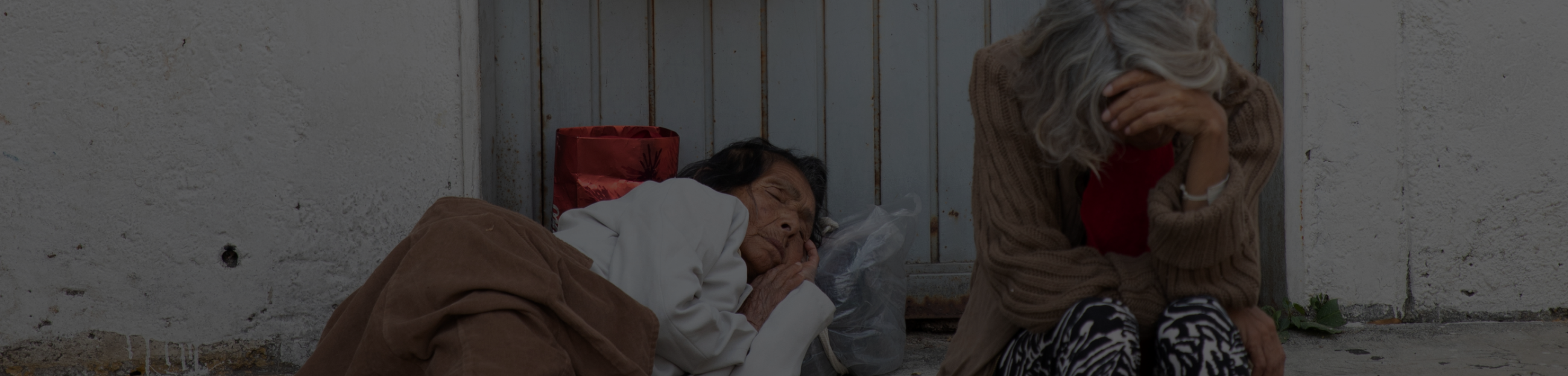 a woman lies on concrete outside, her eyes closed, covered by a thin blanket with her head rested on a plastic bag full of items. Another woman sits next to her with her head hung down resting on her hand.