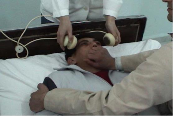 a person being held down by a hand on his shoulder and a hand over his mouth, while another person places a device on his forehead